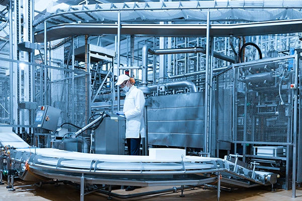 Person in a lab coat and mask working in a large industrial facility with metal machinery.